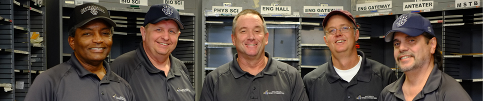 Smiling men with campus unit-labeled mail slots in background